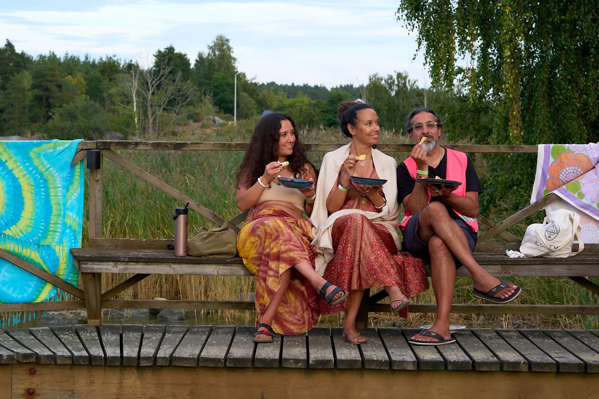Three friends sharing food on a wooden bench beside the water.