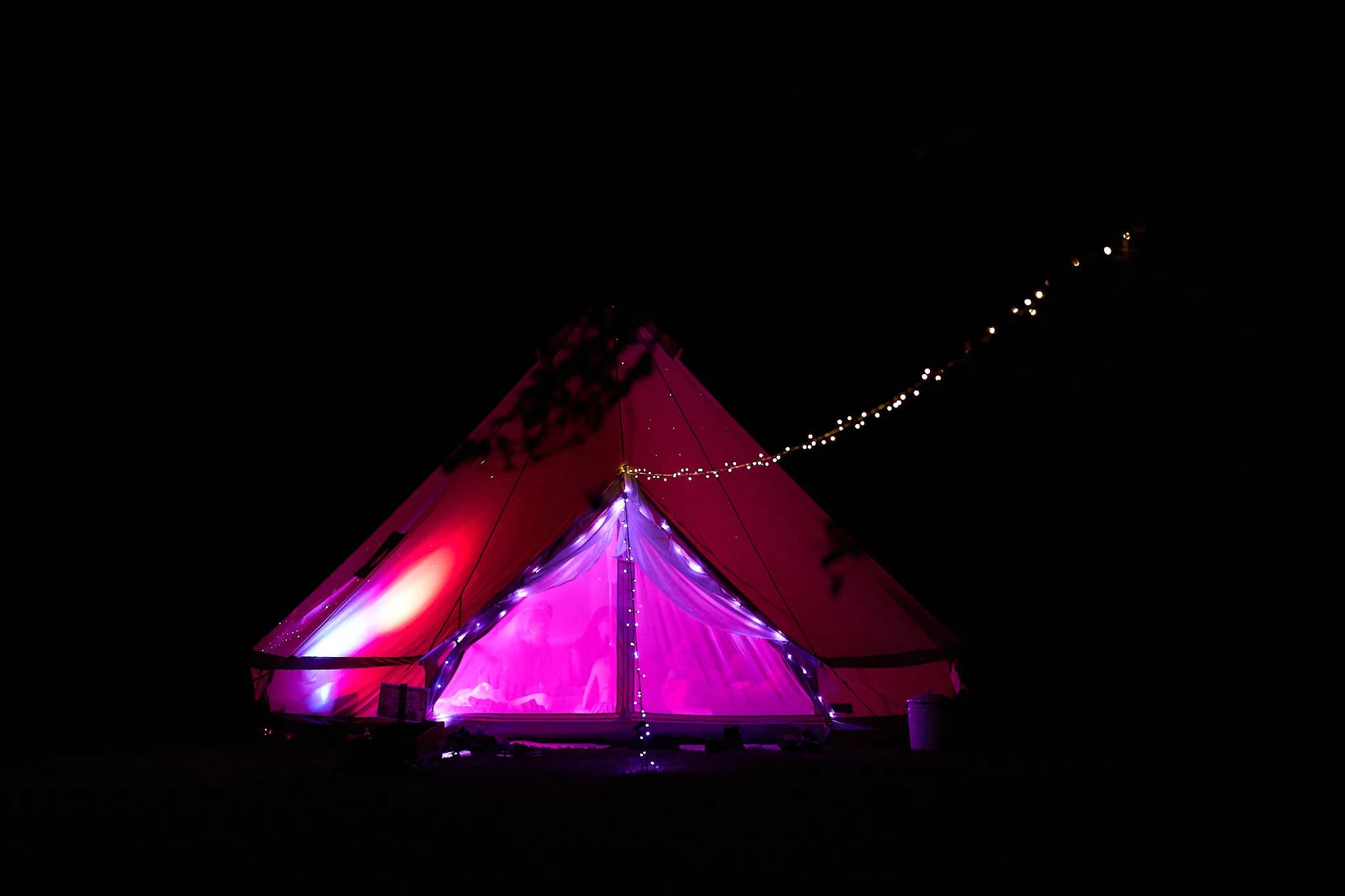 A tipi glowing pink and purple beneath a string of lights at night.