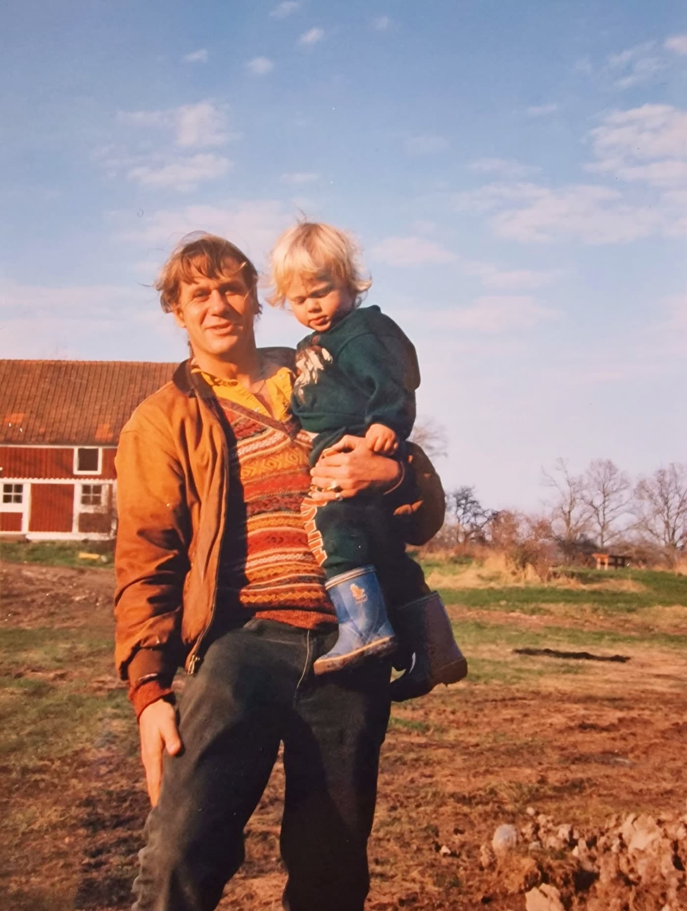 A man carrying a child across the farmyard, the red barn behind