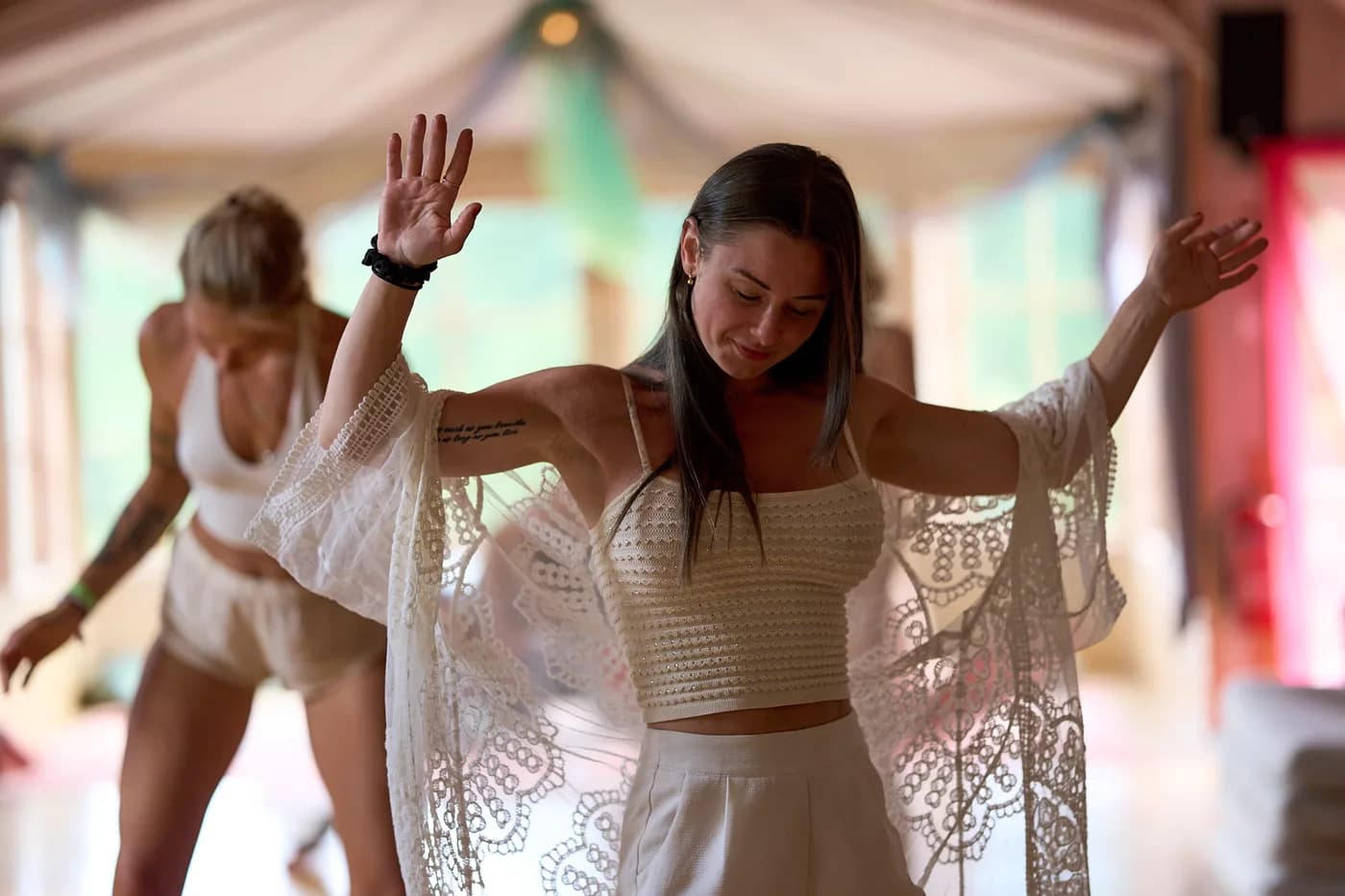 A dancer in a white lace top faces forward with both hands raised.